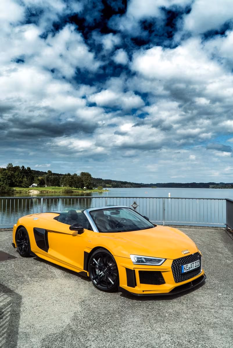 Gelber Audi R8 Spyder vor einem See unter bewölktem Himmel.