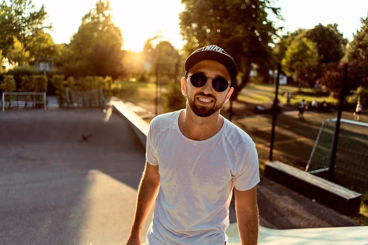 Lifestyle: Lächelnder Mann mit Kappe und Sonnenbrille im Skatepark bei Sonnenuntergang.