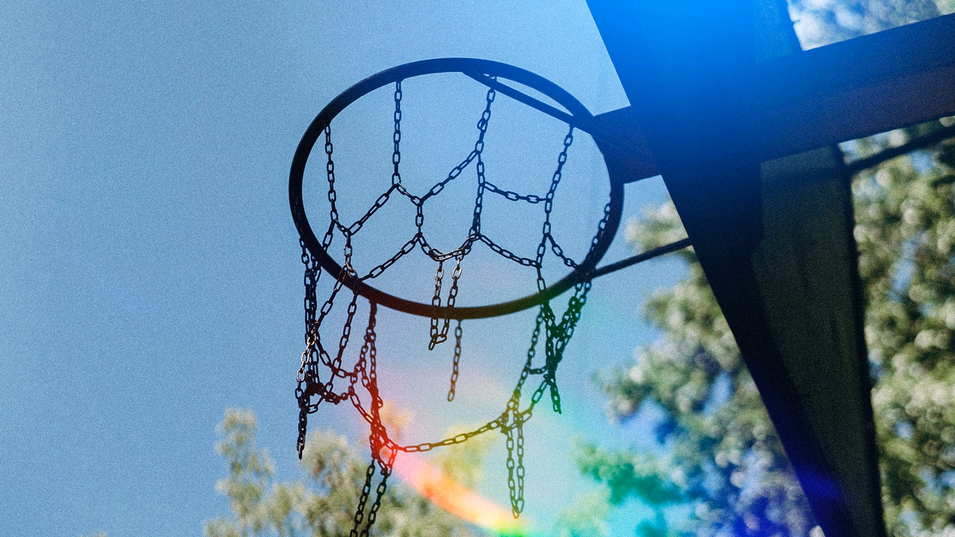 Basketballkorb mit Kettennetz, Regenbogenlicht und blauem Himmel im Hintergrund.