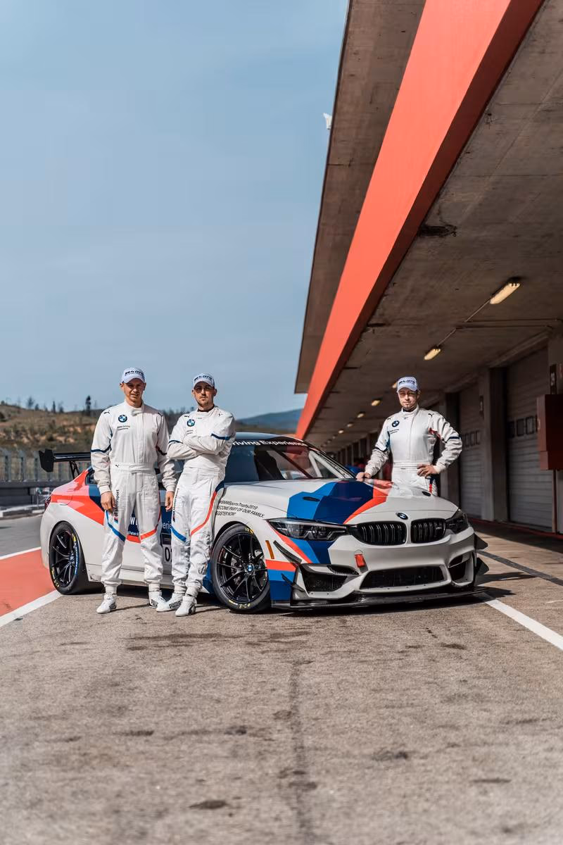BMW Motorsport: Drei BMW-Rennfahrer in weißen Overalls neben einem BMW M4 GT3 im Boxenbereich.