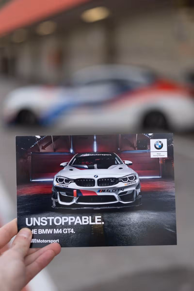 Hand hält BMW M4 GT4 Motorsport-Druck mit Slogan „UNSTOPPABLE“ vor unscharfem Rennwagen.
