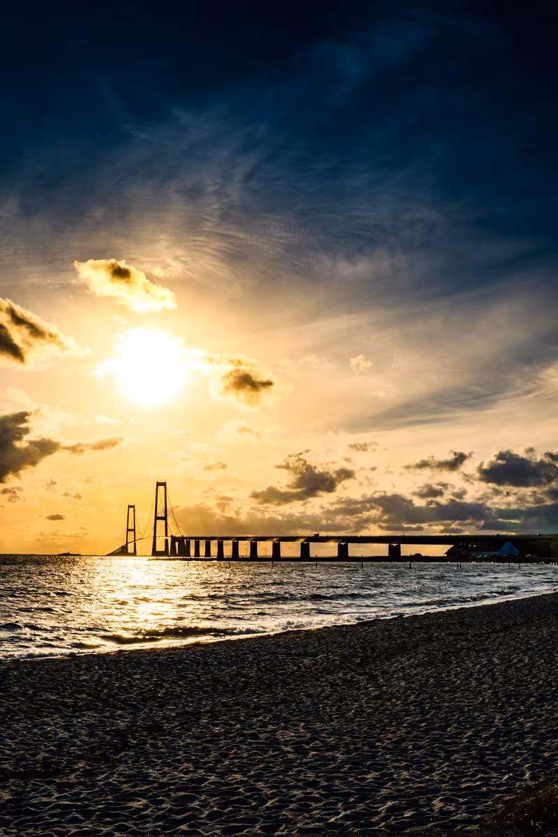 Copenhagen: Silhouettierter Hängebrücken-Sonnenuntergang über dem Meer mit Strand im Vordergrund.