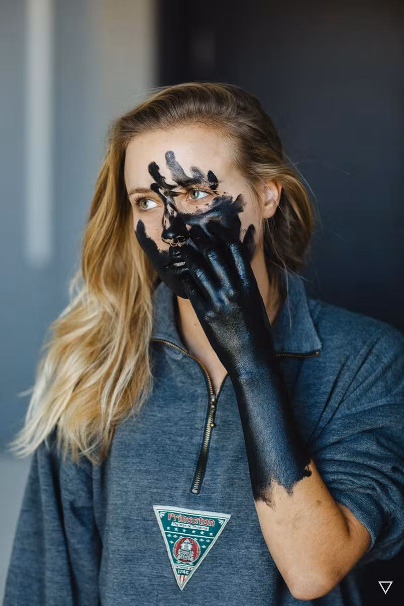 Lifestyle: Frau mit schwarzem Farbanstrich auf Gesicht und Hand vor grauem Hintergrund.