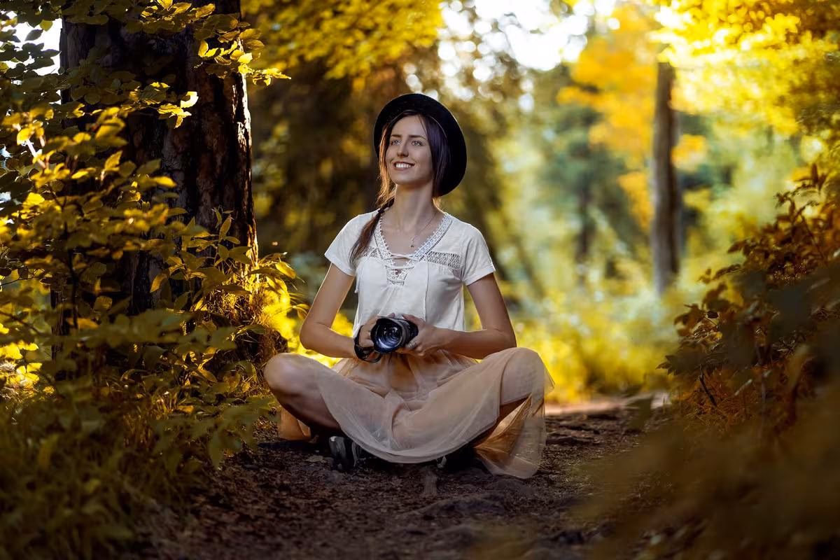 Lifestyle: Frau mit Hut und Kamera sitzt lächelnd auf einem Waldweg im Herbst.