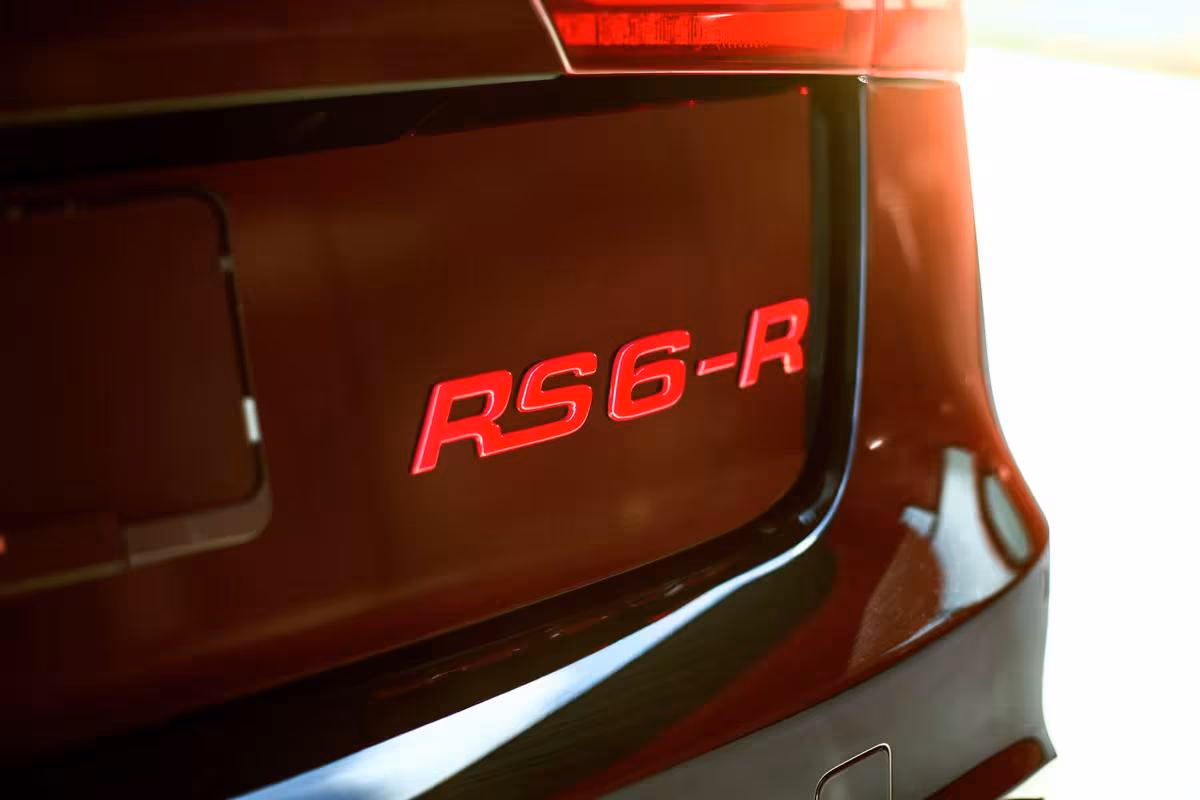 Audi: Rotes RS6-R-Emblem auf der Kofferraumklappe eines dunklen Autos.