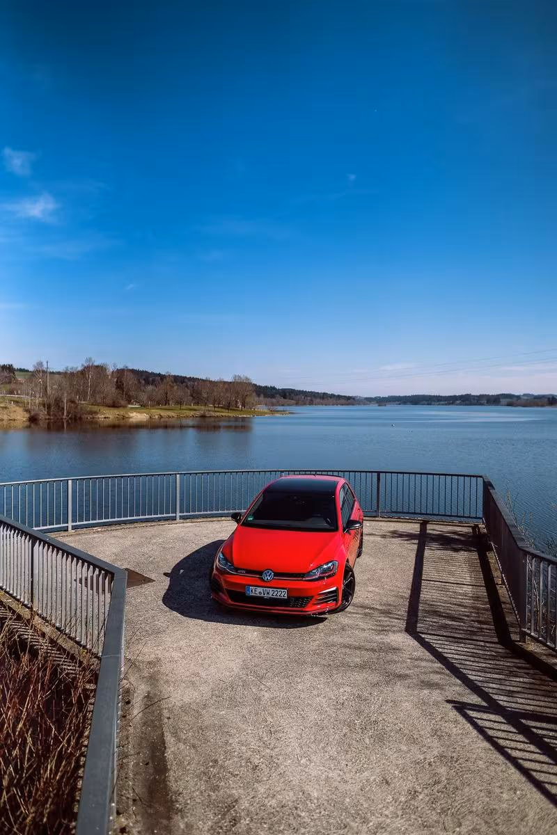 Volkswagen: Roter VW Golf GTI auf einer Betonplattform vor einem See unter blauem Himmel.