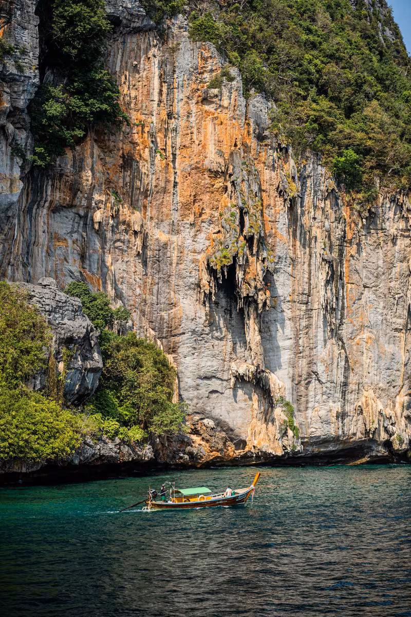 Thailand: Langschwimmboot vor steiler, bewachsener Kalksteinfelswand auf türkisfarbenem Wasser.