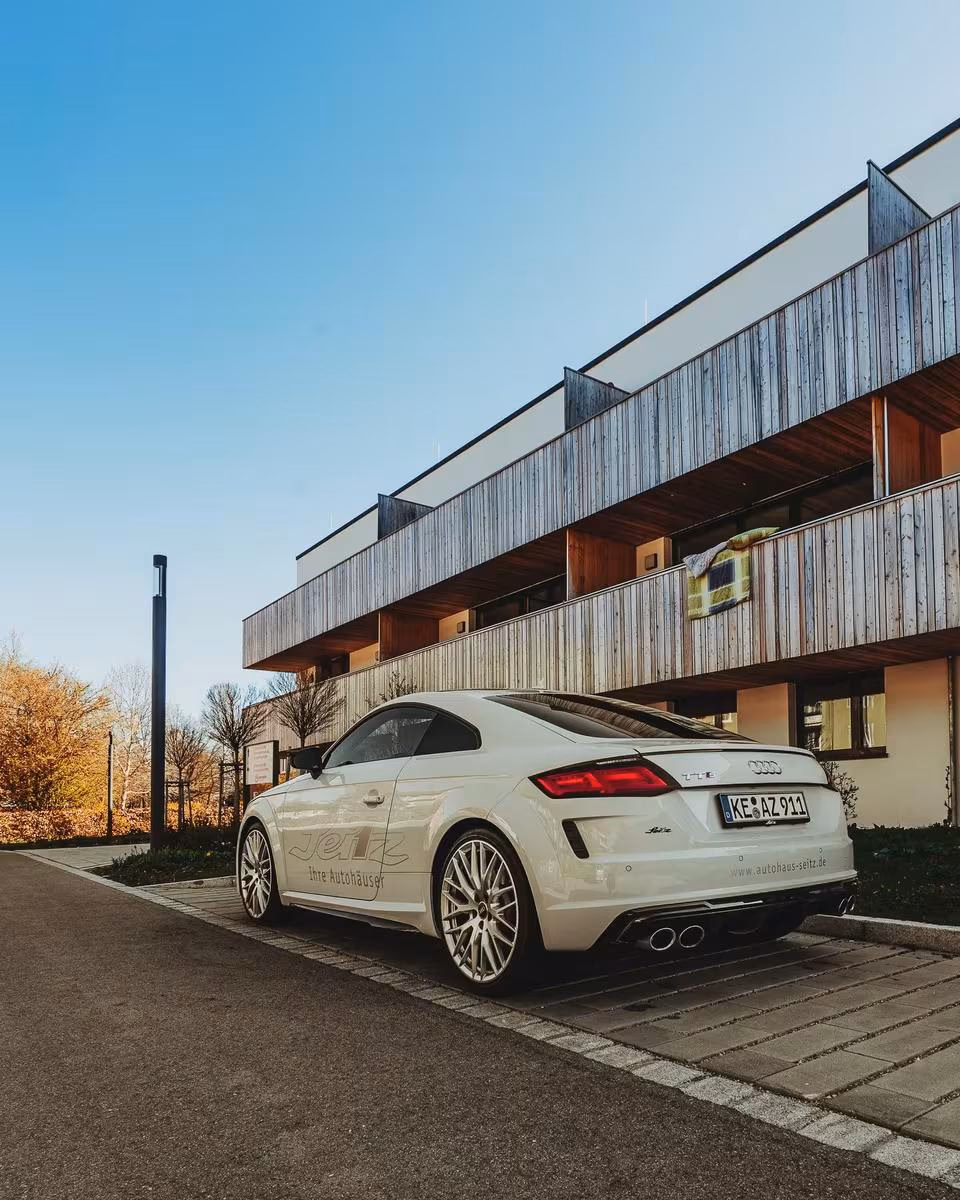 Weißer Audi TT vor modernem Holzgebäude mit Aufkleber „Jetz Ihre Autohäuser“.