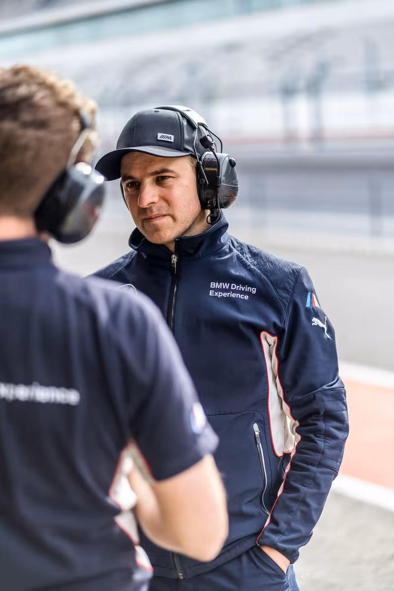 Mann mit Kopfhörer und BMW Driving Experience Jacke auf einer Rennstrecke.