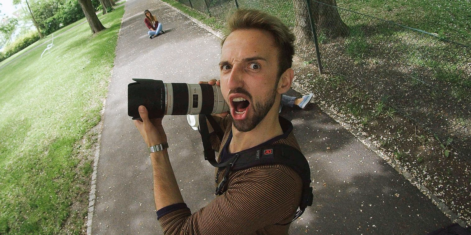 Fotograf hält eine Kamera mit Teleobjektiv und zieht eine übertriebene, komische Grimasse.