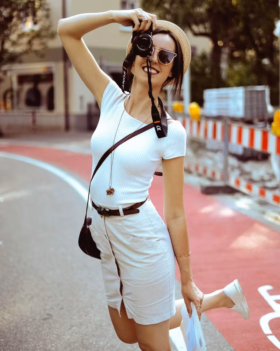 Lifestyle: Frau mit Kamera, Sonnenbrille und Hut lächelt auf einer Stadtstraße.