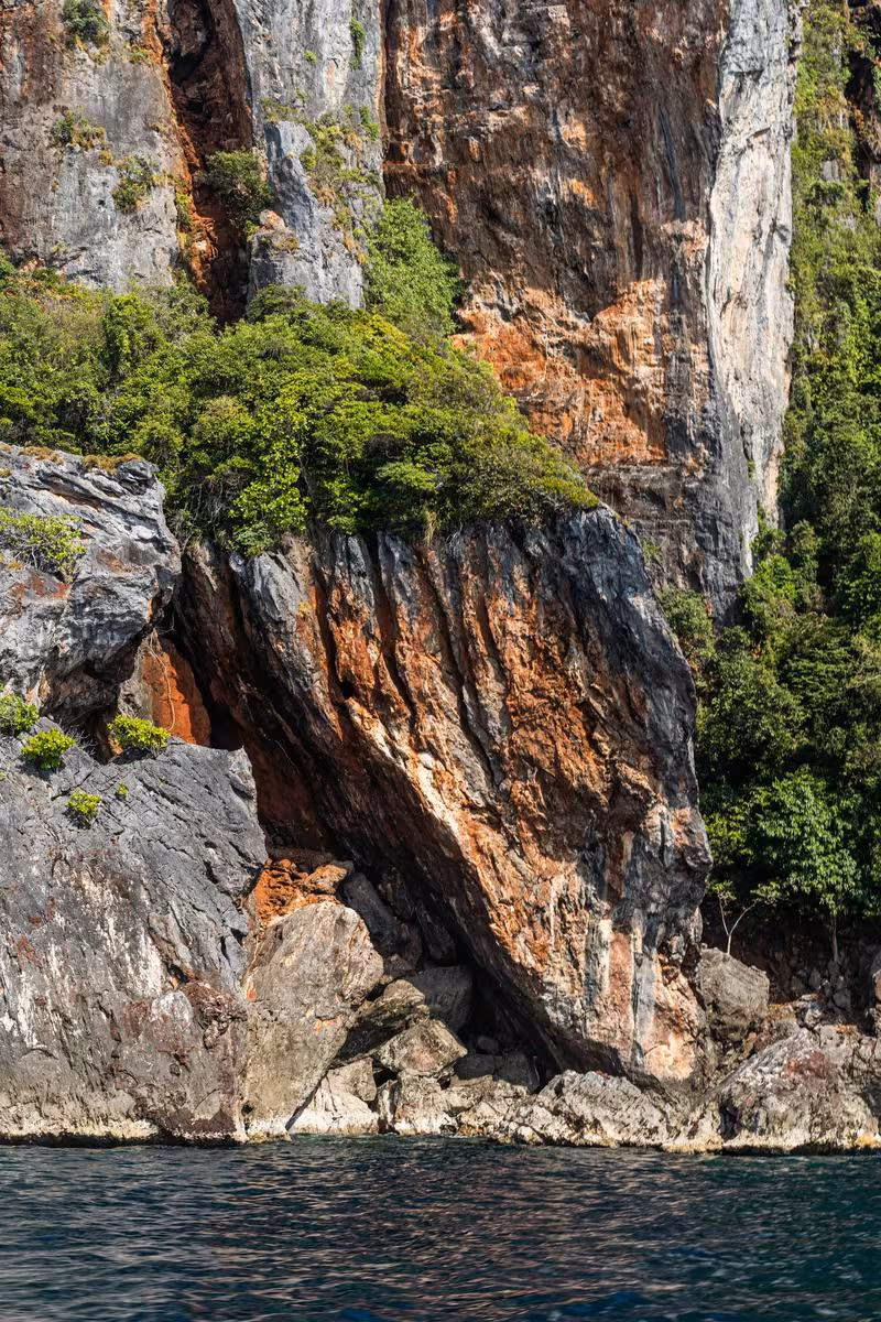 Thailand: Steile, bewachsene Klippe mit rostfarbenen Felsformationen am Meer.