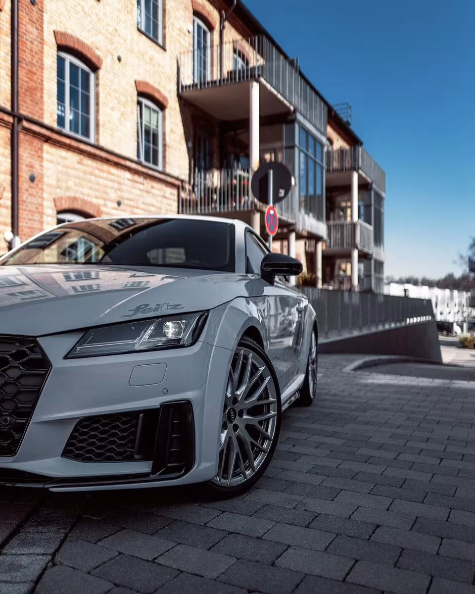 Grauer Audi mit „Feiz“-Schriftzug vor einem Backsteinhaus auf Kopfsteinpflaster.