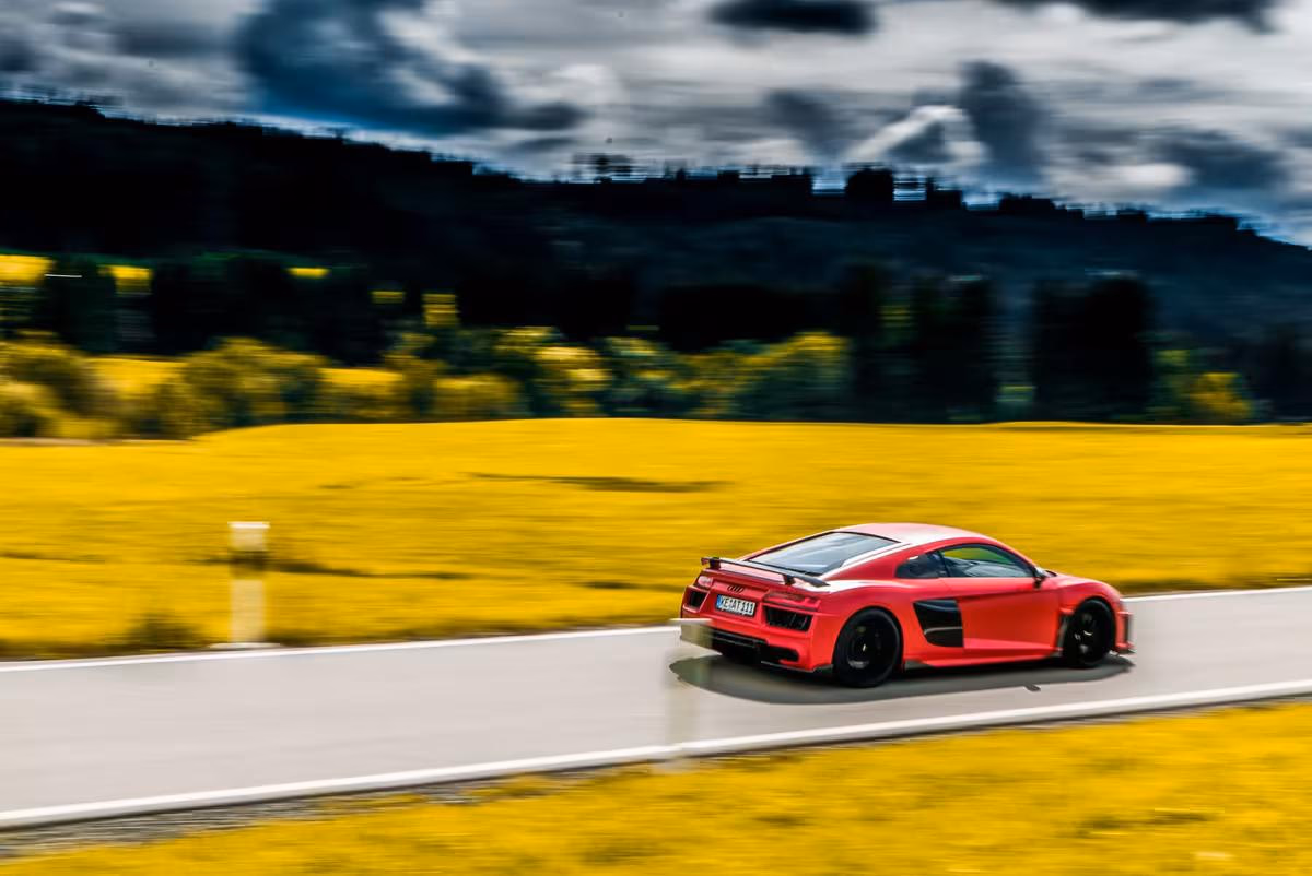 Audi: Rotes Sportcoupé fährt auf Landstraße durch gelbes Feld bei bewölktem Himmel.