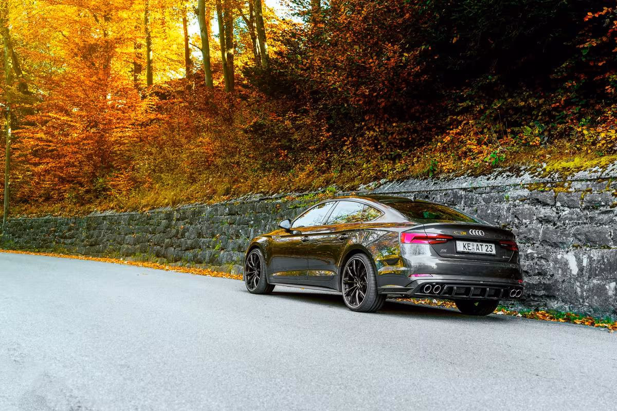 Schwarzer Audi RS7 Sportback auf Landstraße vor herbstlichem Wald und Steinmauer.