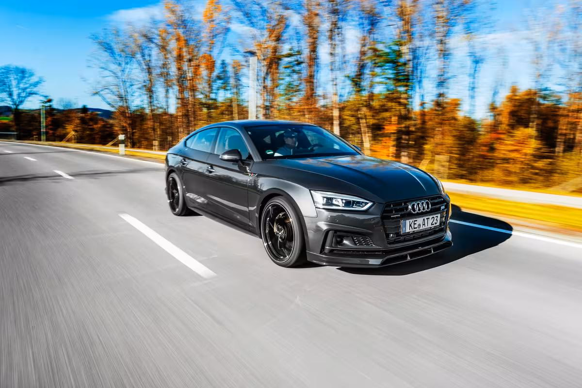Schwarzer Audi RS 5 Sportback fährt auf einer Landstraße durch eine herbstliche Waldlandschaft.